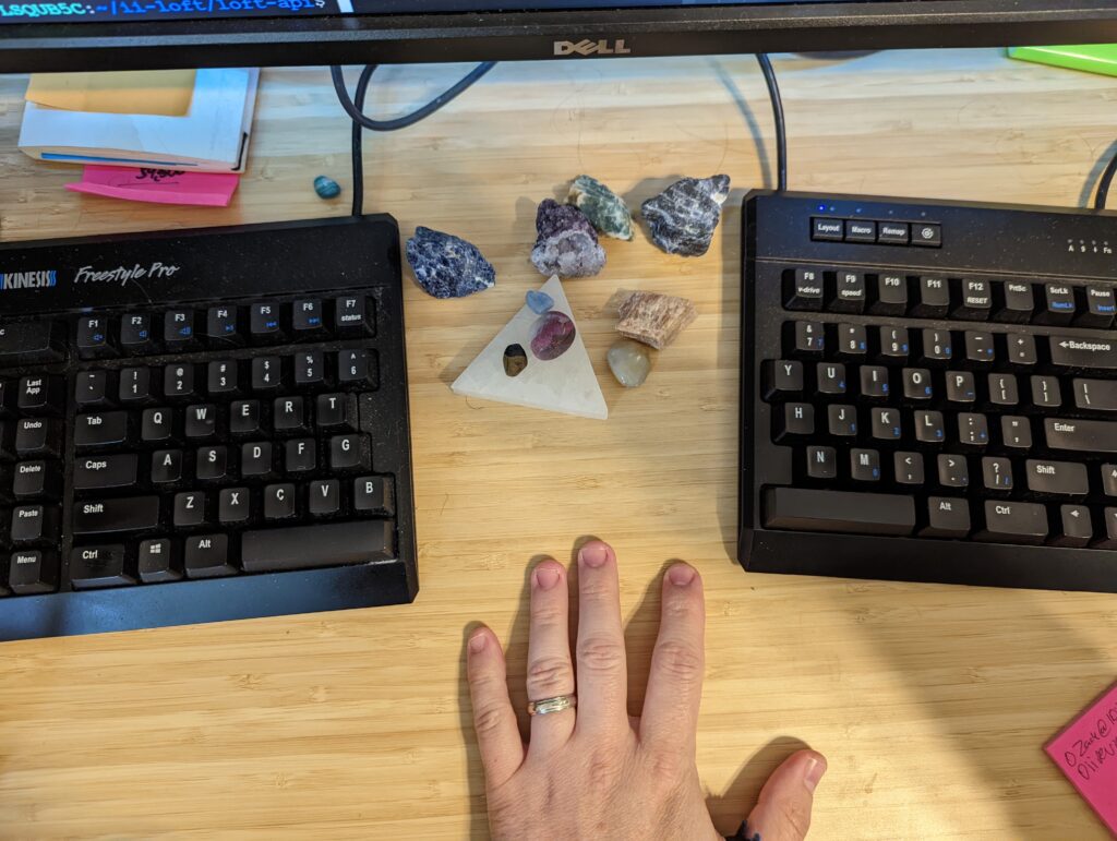 Split Keyboard in front of computer screen,  rocks arrayed on a crystal triangle under a monitor. A hand is in between keyboard parts