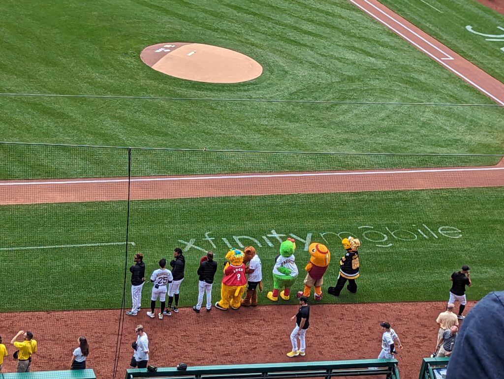 Several sports mascots, Pittsburgh Parrot, Pittsburgh Penguin Guy, and some other mascots, along with Pirates players lined up on the baseline.