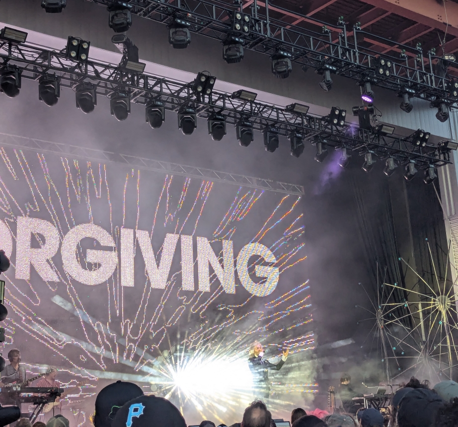 Wayne from Flaming Lips on stage, arms outstretched, in front of the word "Forgiving" and burst of light.