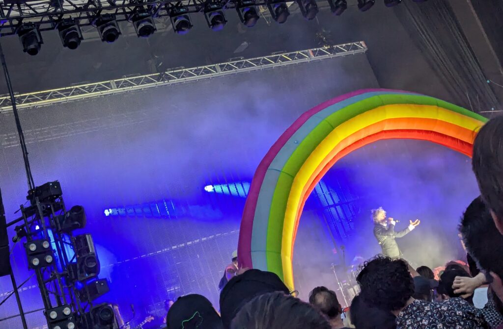 Wayne from Flaming Lips on stage, arms outstretched, under an inflated rainbow